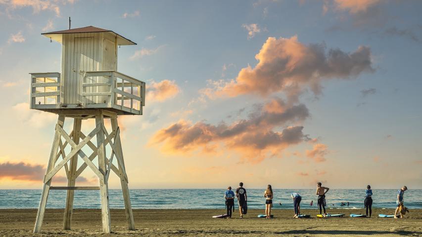 Imagen de Agosto del Calendario 2026 (Manuel Rueda) con una instantánea de un grupo de surfistas equipándose para realizar deporte